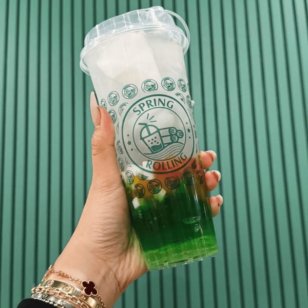 A hand holds a Spring Rolling branded cup with a green drink and colorful jellies against a green wall.